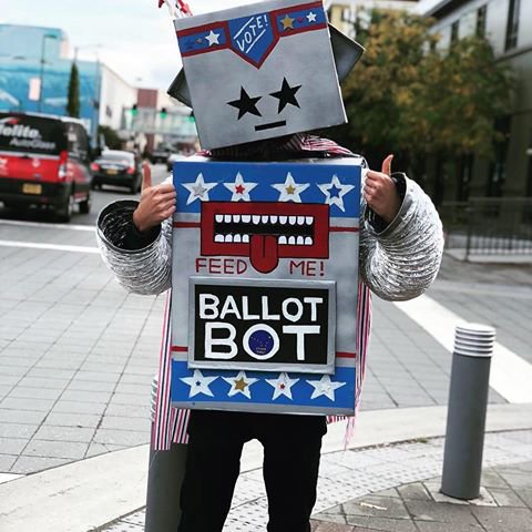 Person giving thumbs up, dressed up as a ballot box with a mouth and the words, "Feed Me. Ballot Box."